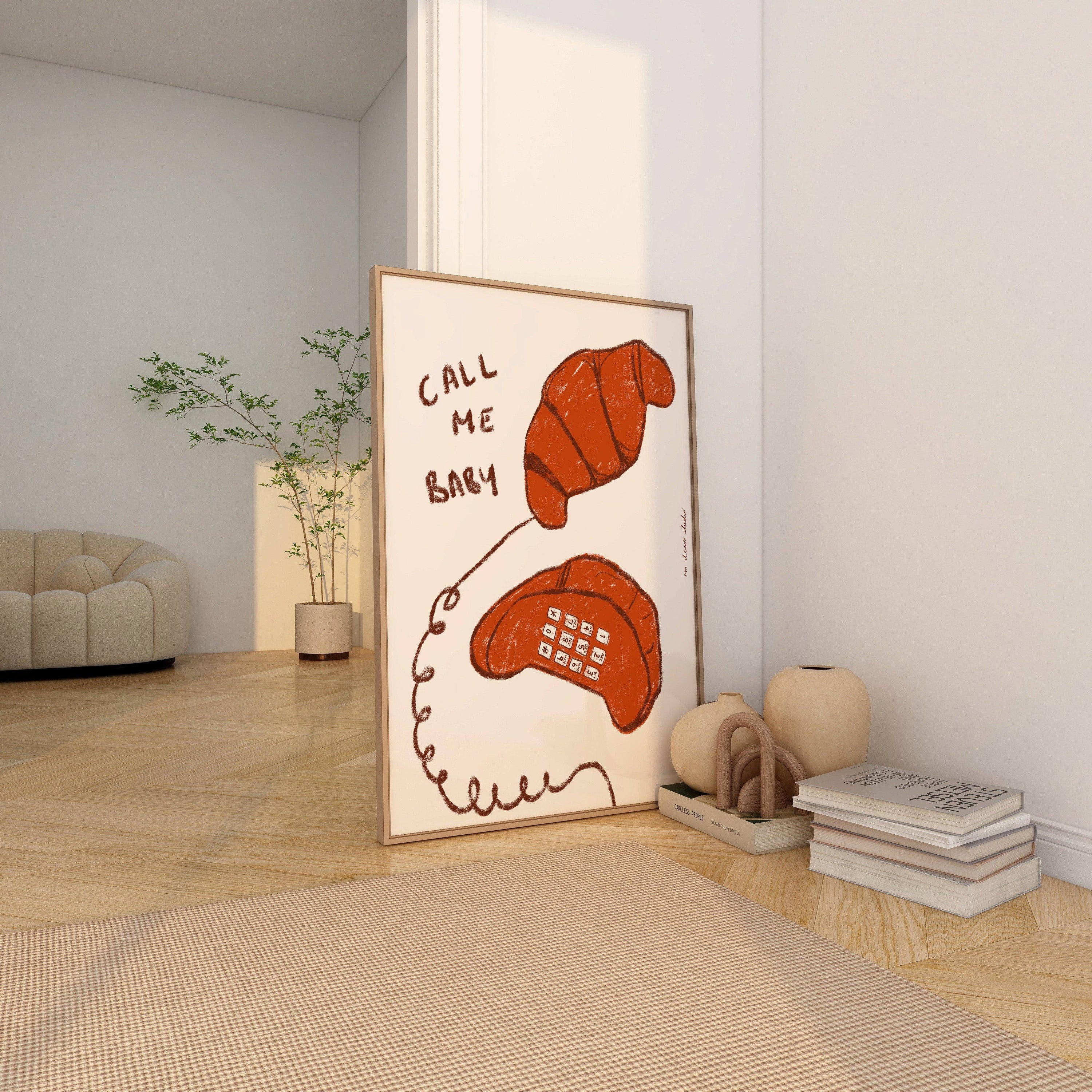 A framed print of a croissant depicted as a phone with the phrase 'Call Me baby' written on it, placed on the floor in a home setting.