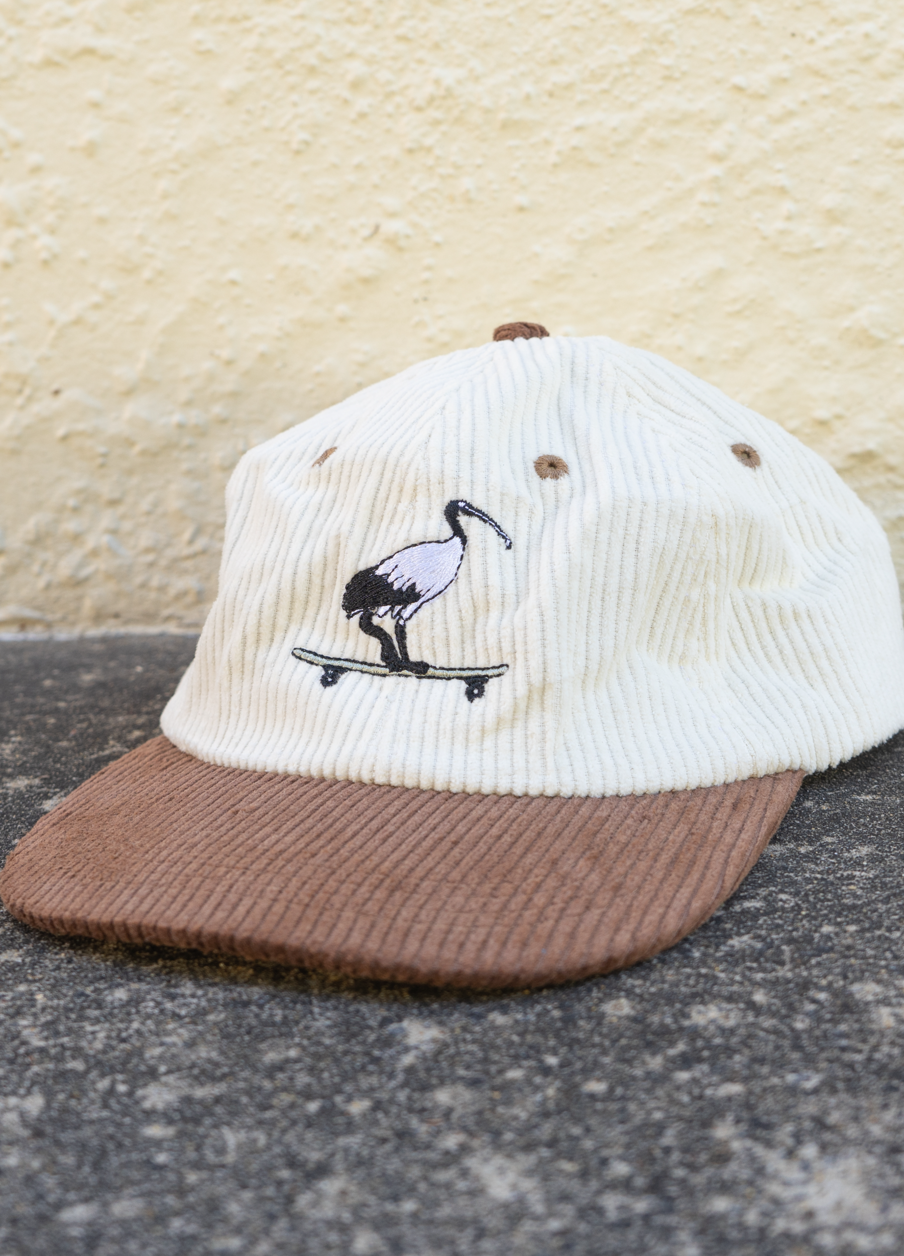 White and brown cap with a stork on a skateboard design against a textured beige background.