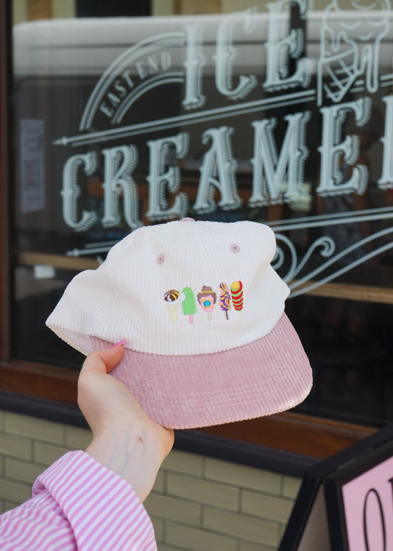 Pink cap with embroidered designs held in front of an ice creamery sign.