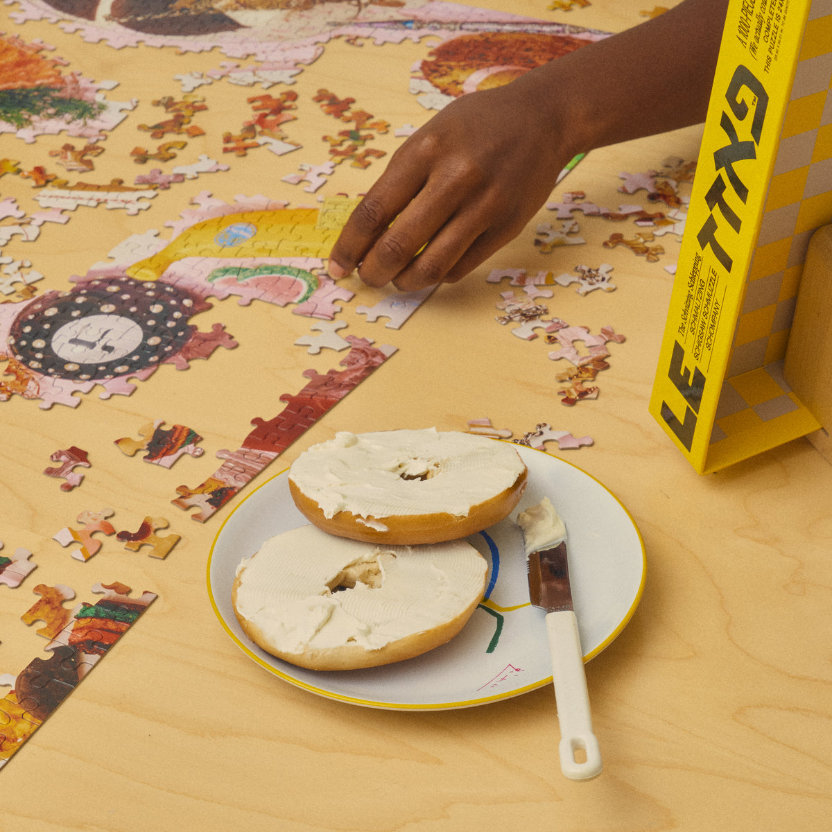 Hand assembling the Jewish American Puzzle beside cream cheese bagels and the open yellow puzzle box.
