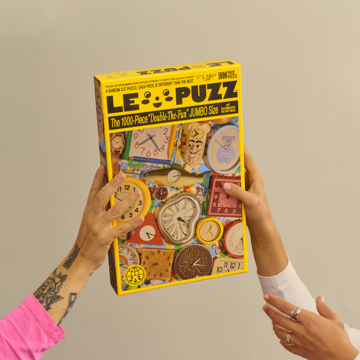 Two people holding the yellow Le Puzz It’s About Time puzzle box featuring a colourful collection of clocks.