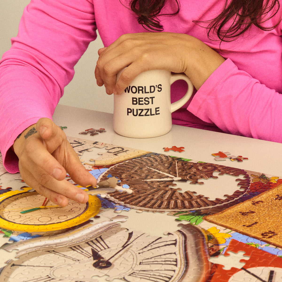 Person assembling the It’s About Time puzzle beside a mug reading “World’s Best Puzzle”.