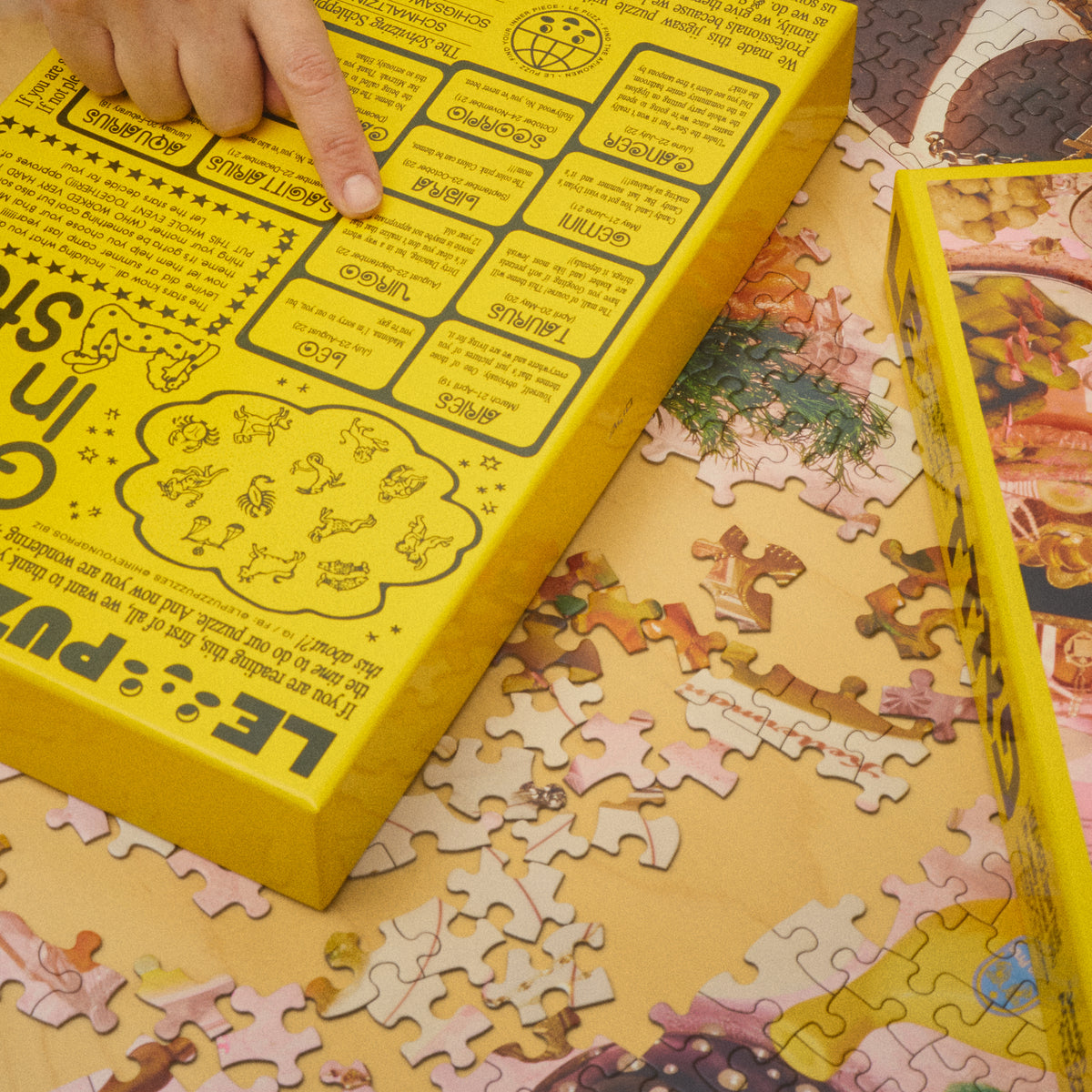 Hand pointing to the zodiac text on the back of the Jewish American Puzzle box, surrounded by jigsaw pieces.