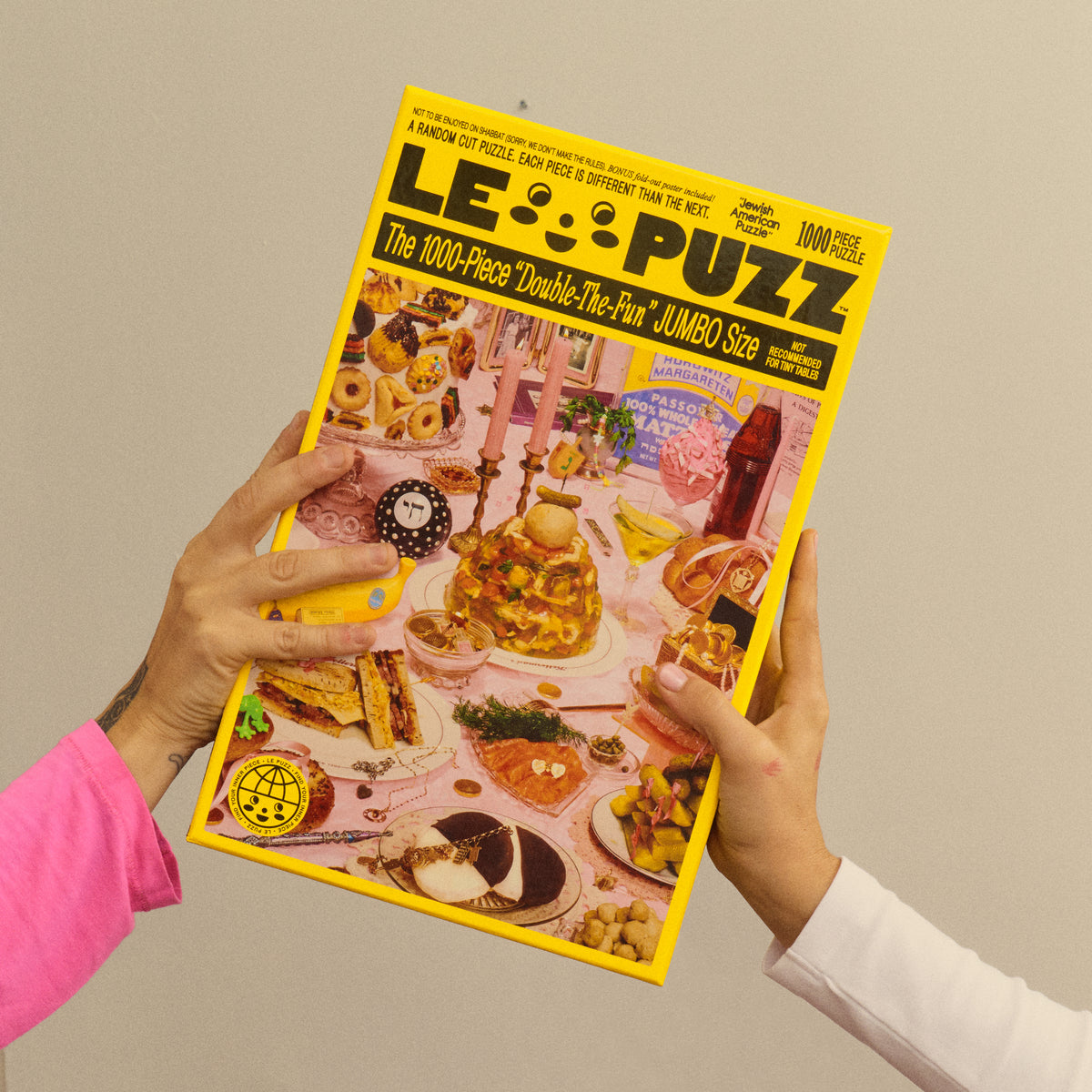 Two people holding the yellow Le Puzz Jewish American Puzzle box featuring a pink table spread of food and candles.