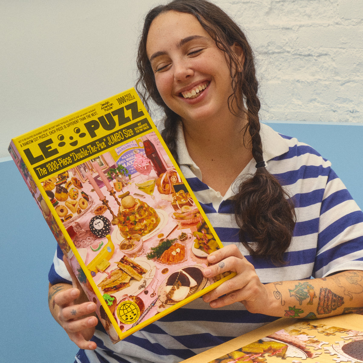 Smiling person in a blue-and-white striped shirt holding the Le Puzz Jewish American Puzzle box.