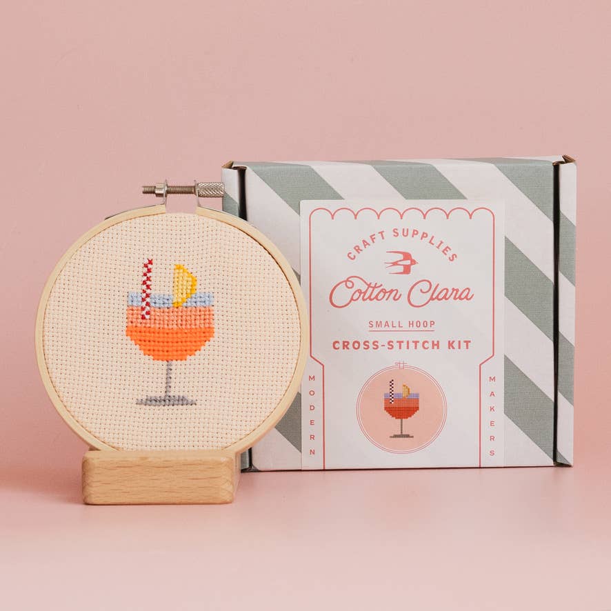 Cross-stitch kit with hoop and completed design next to Cotton Clara packaging on a pink background