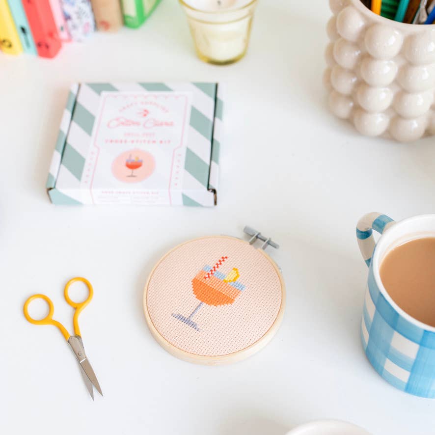 Cross-stitched hoop with a colorful design on a table with scissors, a mug, and other items.