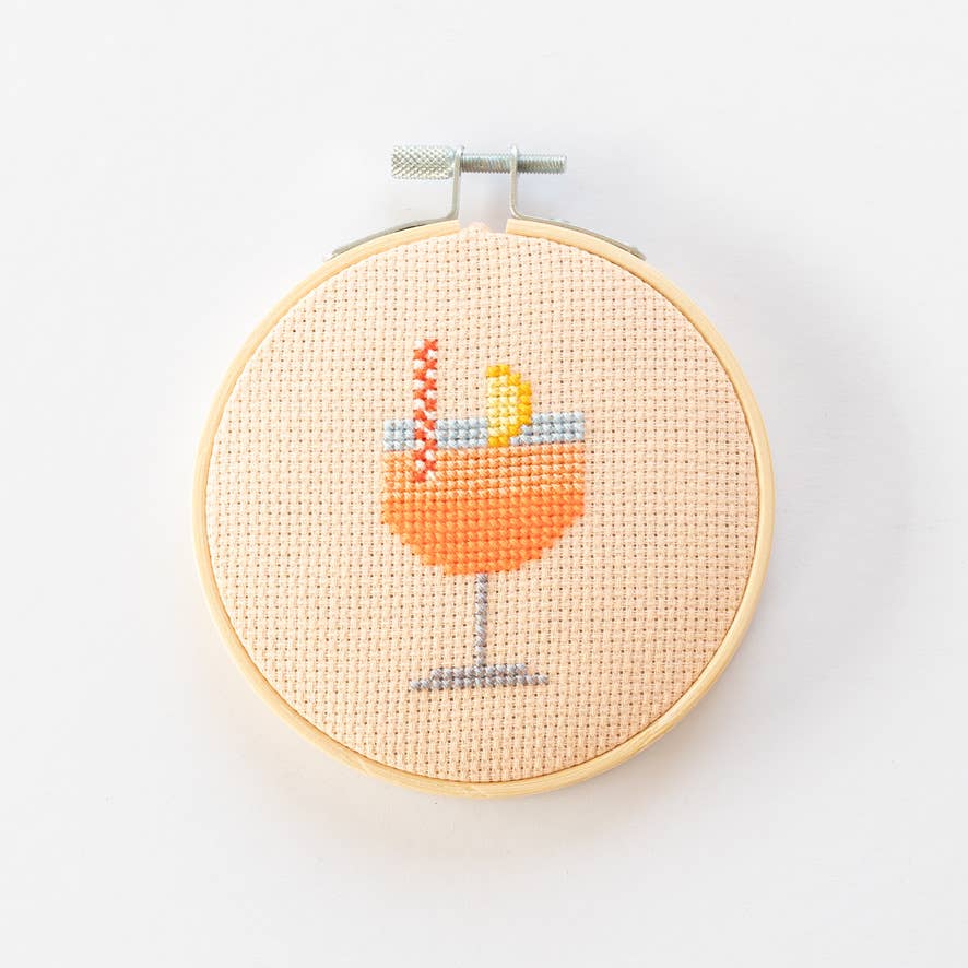 Cross-stitched design of a cocktail glass with a lemon wedge in a wooden hoop on a white background