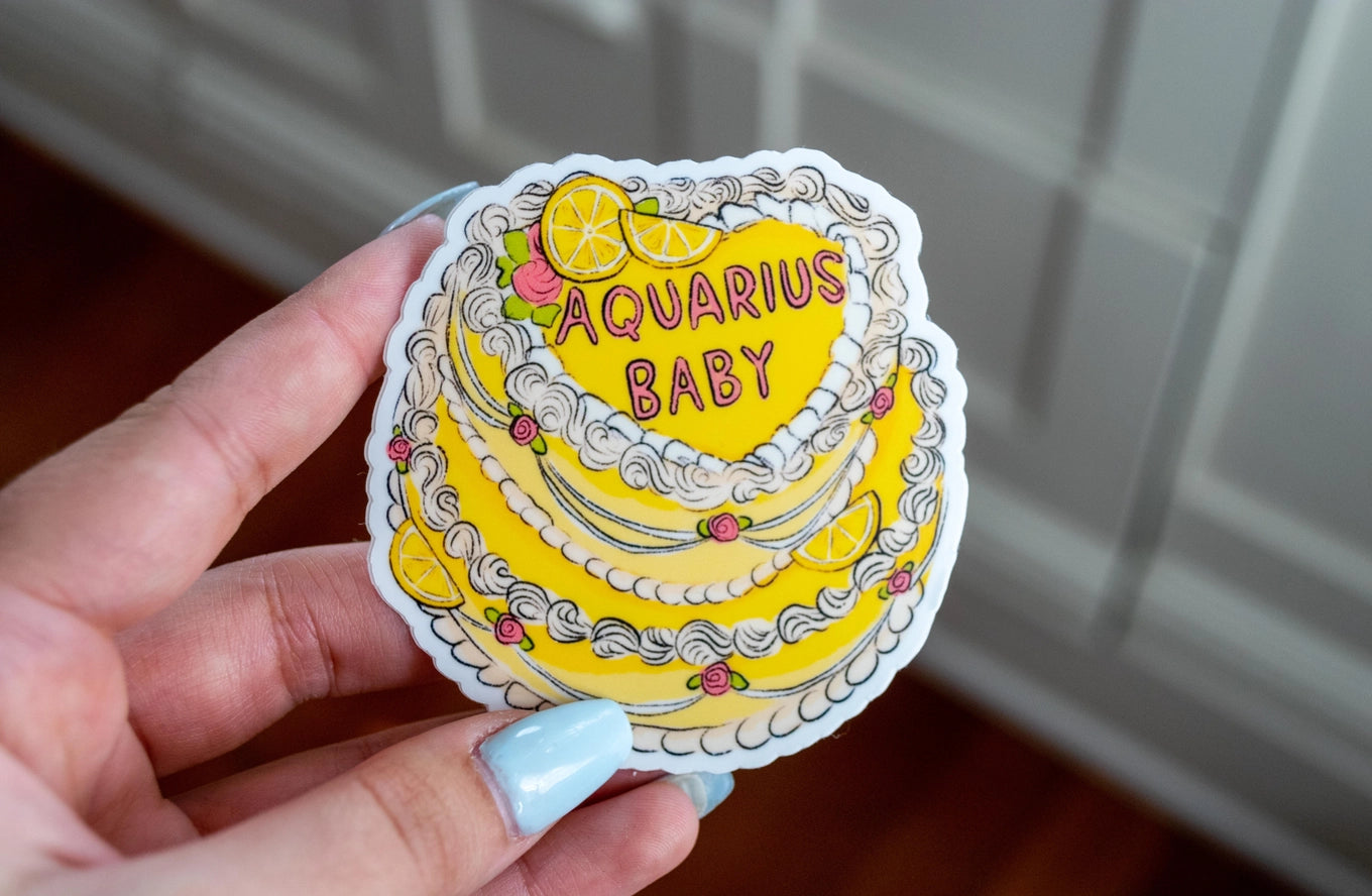 Sticker of a yellow cake with 'Aquarius Baby' text held by a hand.