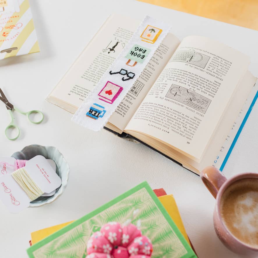 Open book with cross-stitch patterns, a cup of coffee, and stationery items on a table.