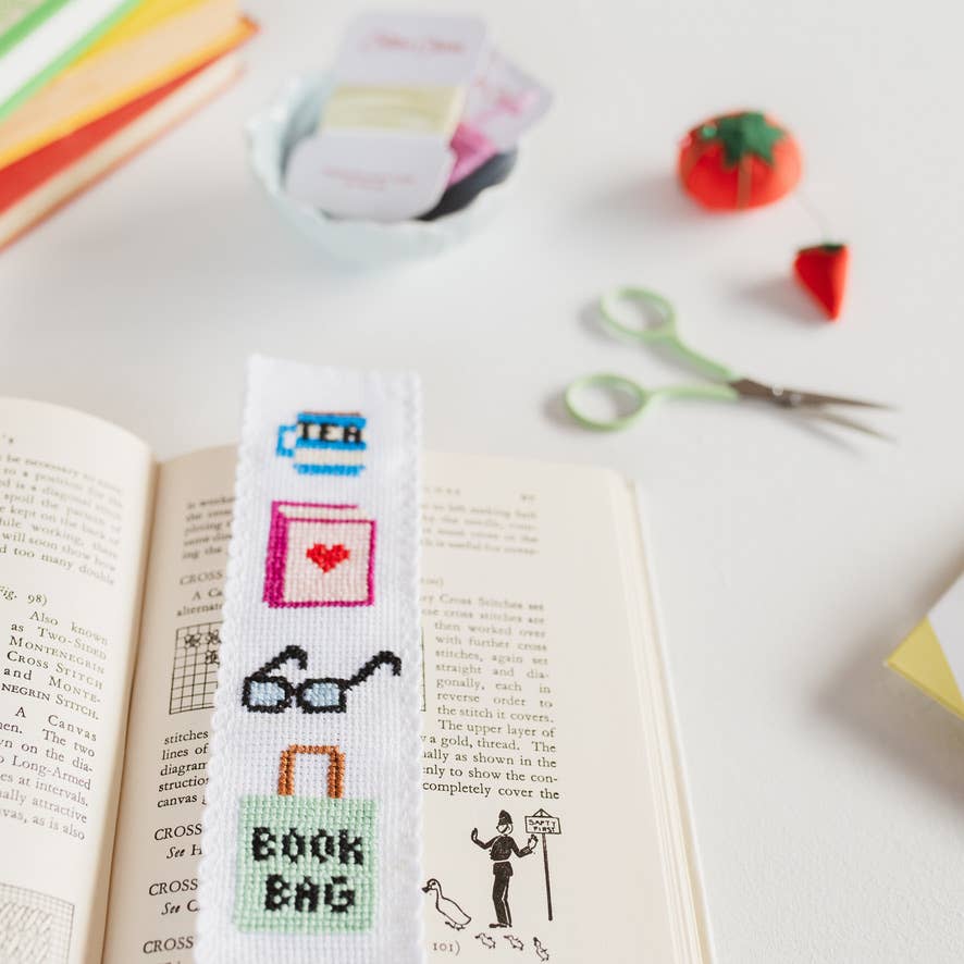 Cross-stitched bookmarks on an open book with stationery items in the background