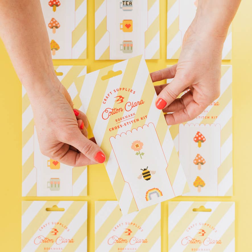 Hands holding a Cotton Clara cross-stitch kit against a yellow background with patterned cards.