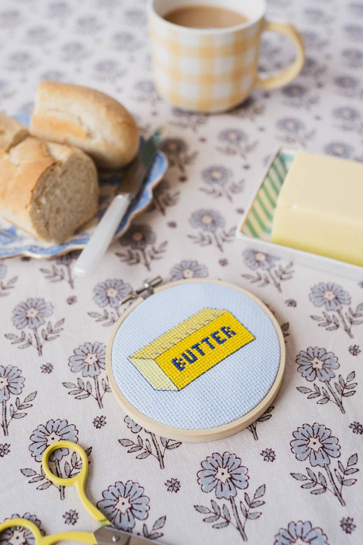 Embroidery hoop with a 'butter' design on a floral tablecloth with bread and coffee.