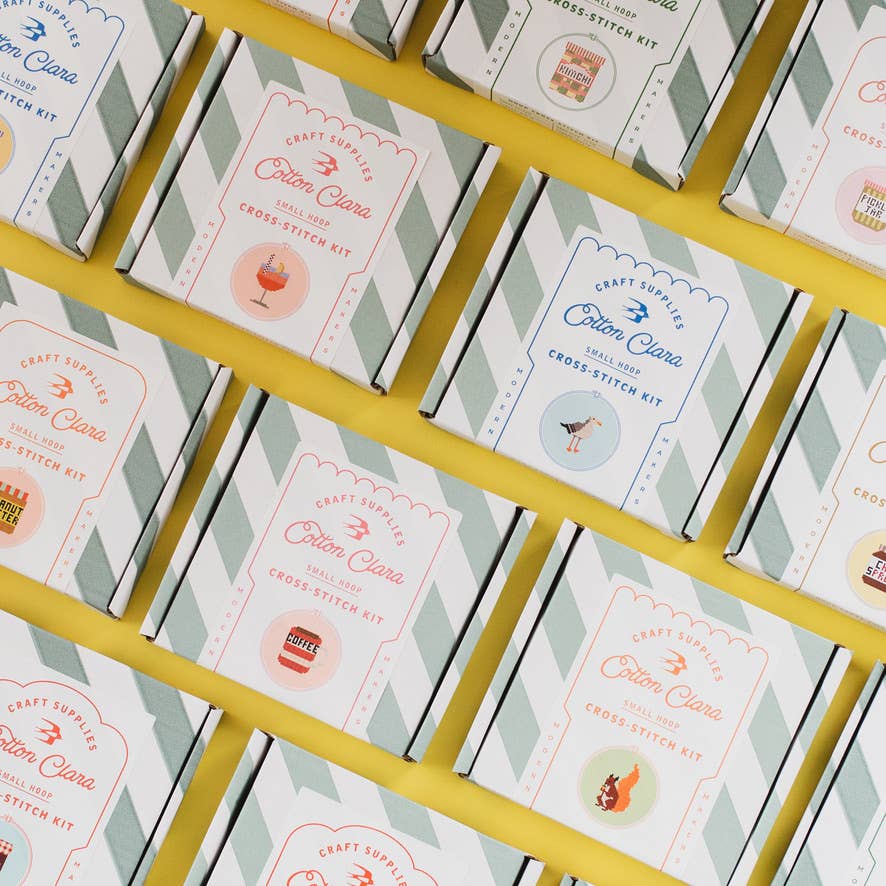 Grid of Cotton Clara cross-stitch kit boxes on a yellow and white striped background