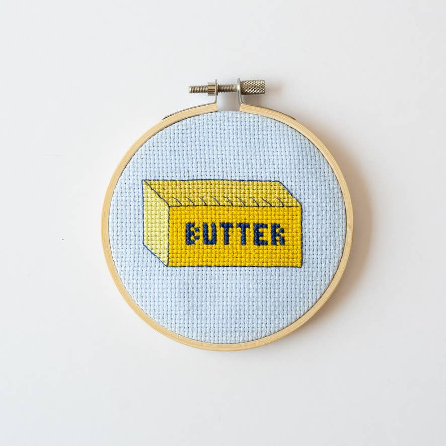 Embroidery hoop with a cross-stitched design of a butter container on a light blue background.