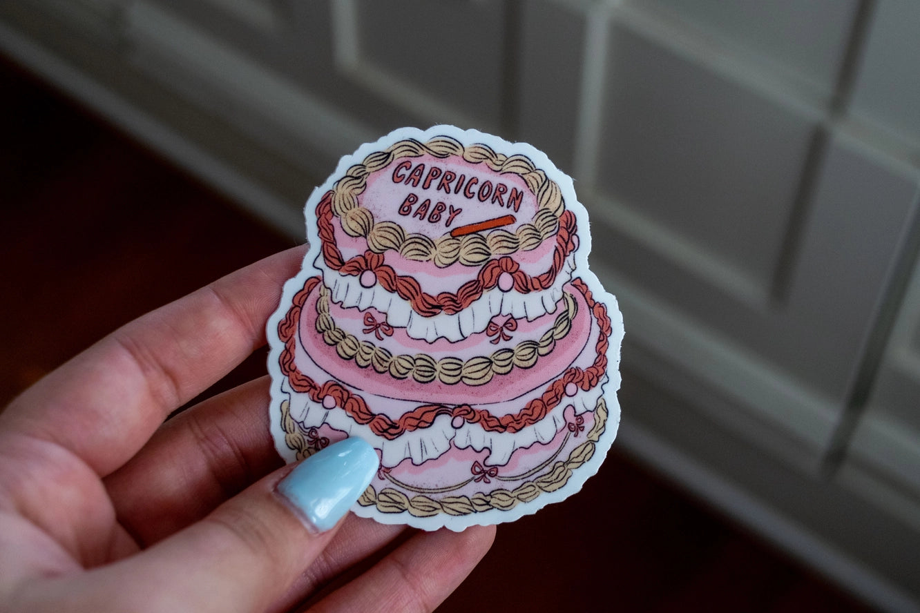 Sticker of a three-tiered cake with 'Capricorn Baby' text held in a hand.