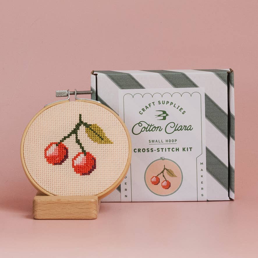 Cross-stitch kit with cherry design and Cotton Clara packaging on a pink background