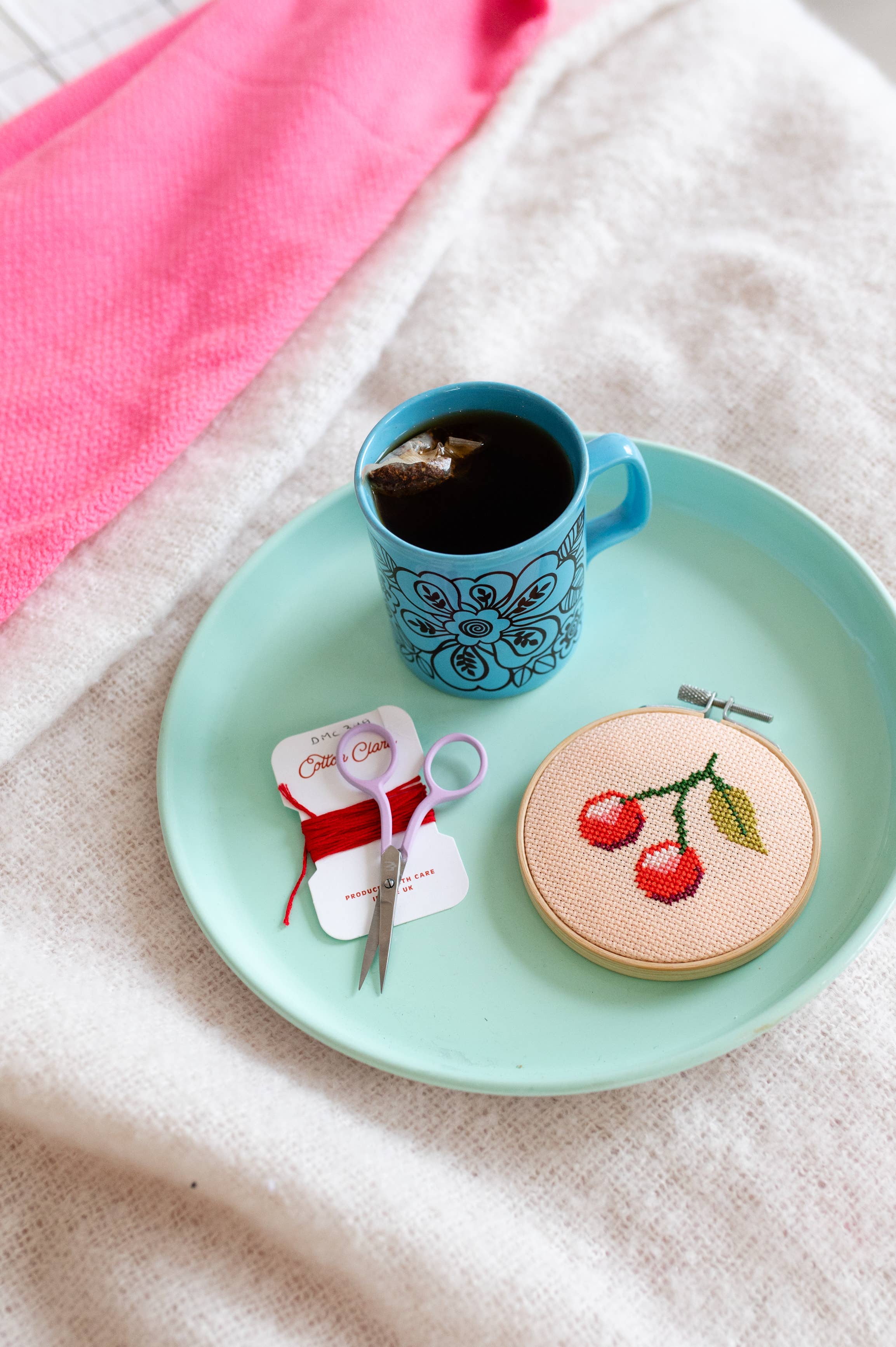 Teal plate with a blue mug, embroidery hoop, and scissors on a textured surface.