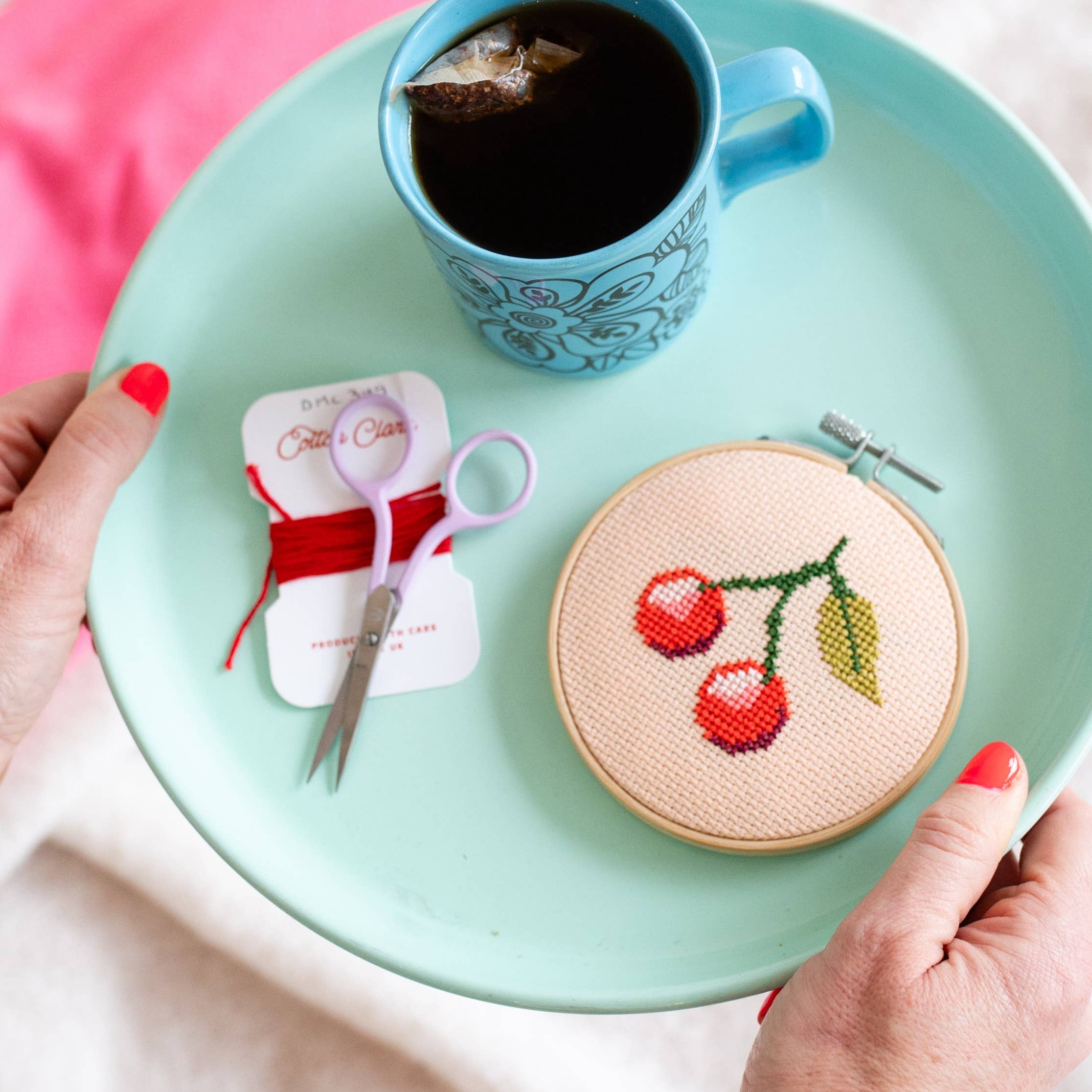 Cross-stitched cherry design on a wooden hoop with scissors, thread, and a cup of tea on a green tray.