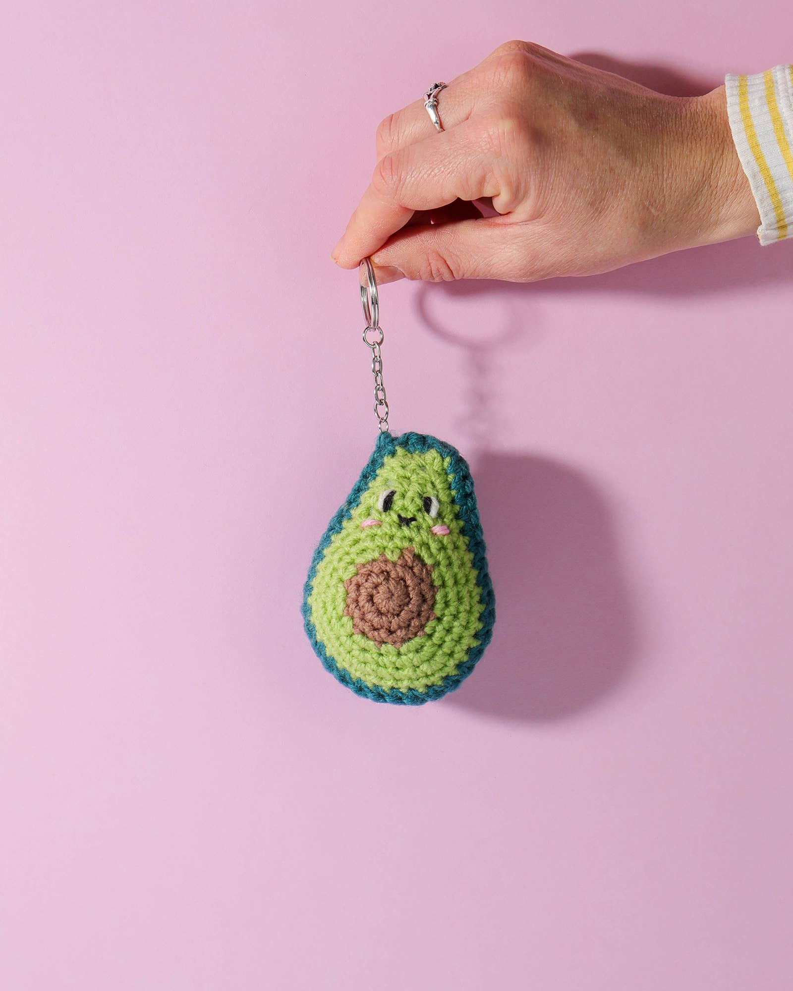Hand holding a crocheted avocado keychain against a pink background