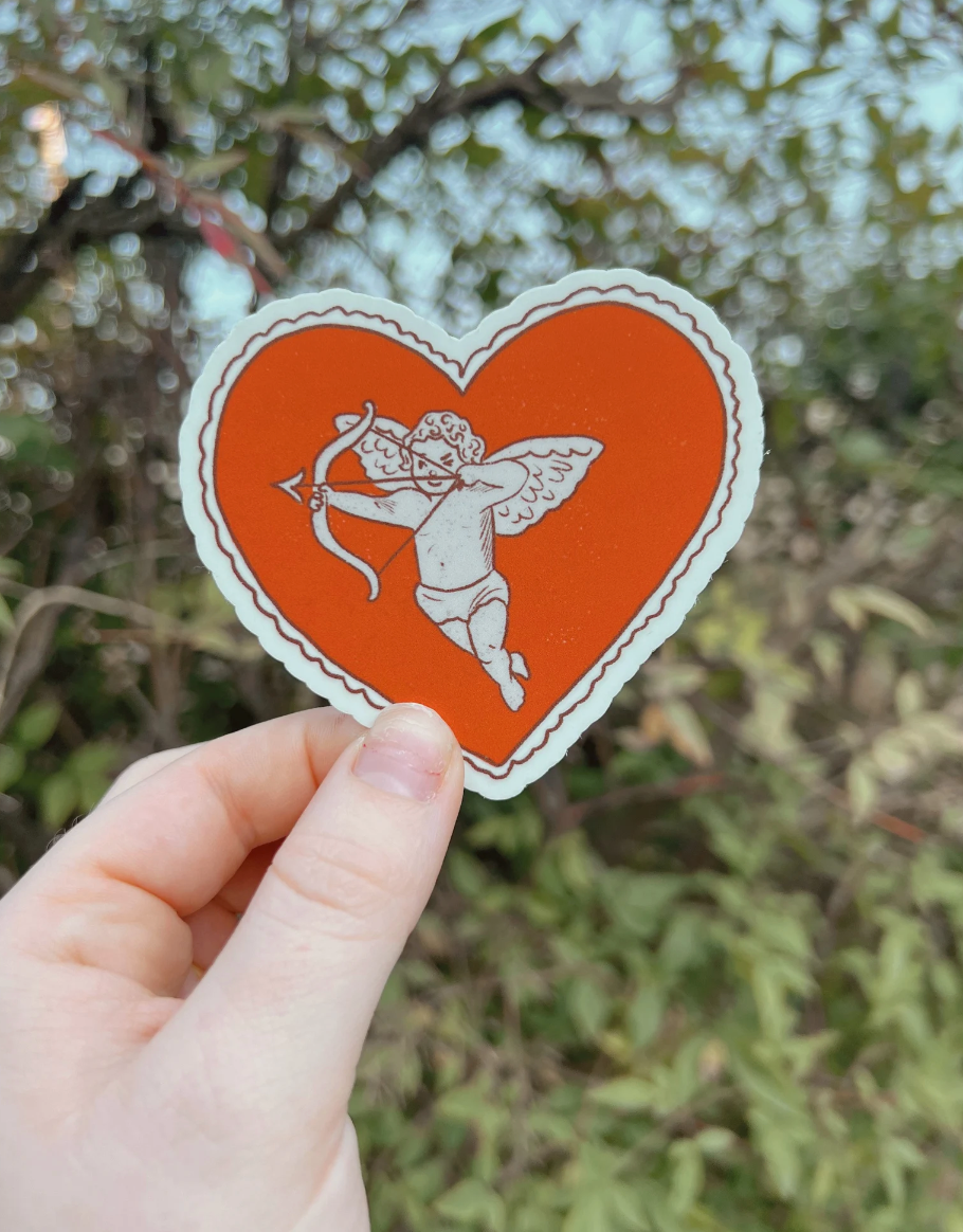 Heart-shaped sticker with Cupid design held by a hand against a blurred natural background
