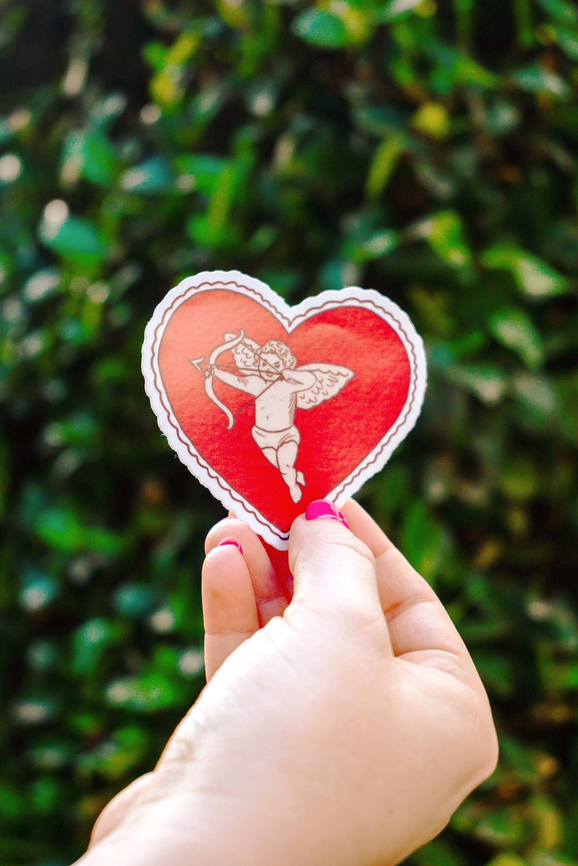 Heart-shaped sticker with a fairy design held by a hand against a green leafy background