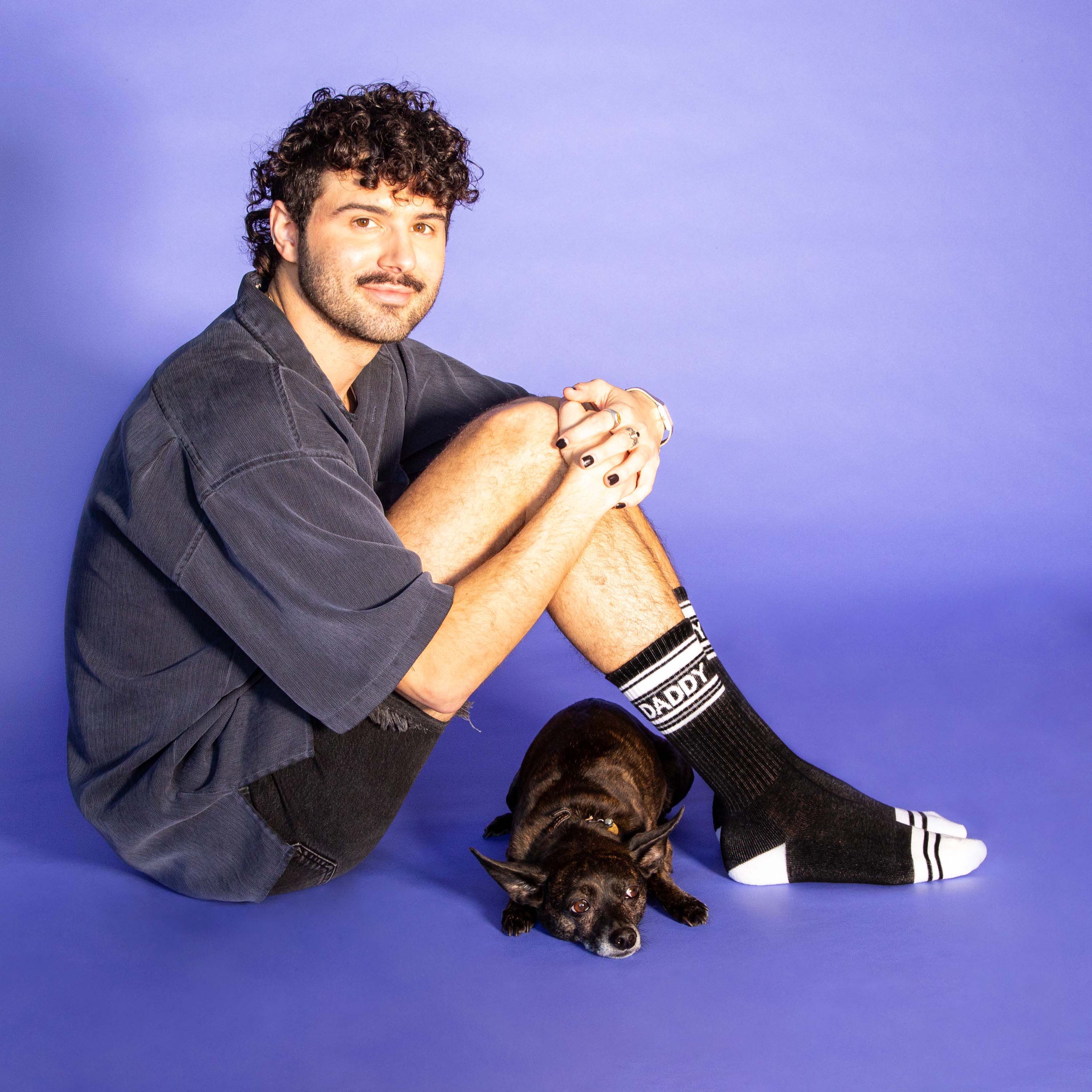 Man sitting on a blue background with a dog, wearing socks with text.