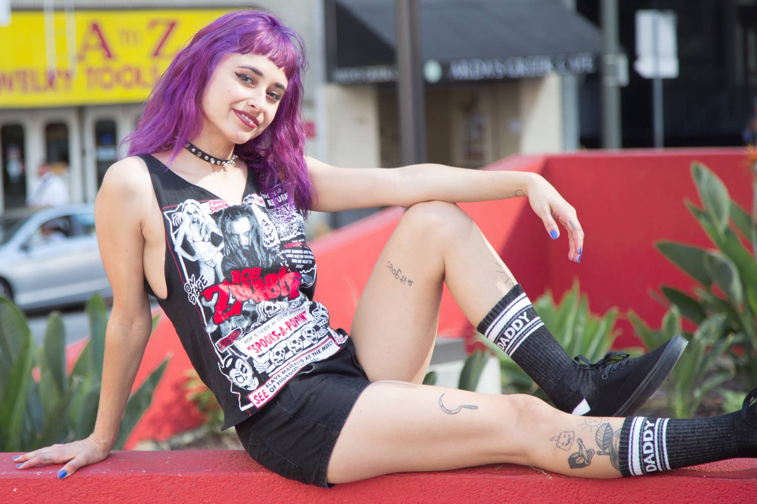 Person with purple hair and tattoos sitting on a red surface outdoors.