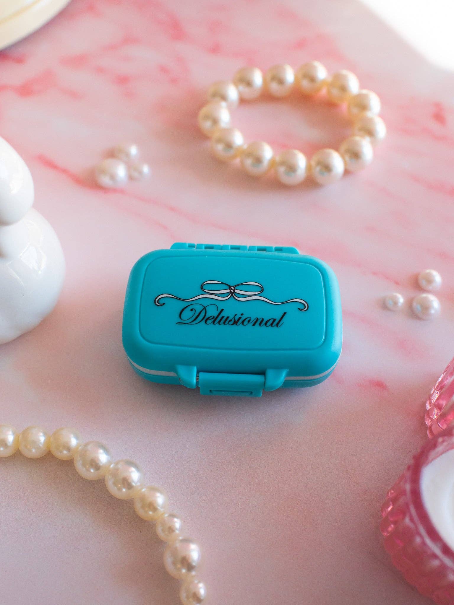 Turquoise pill box with 'Delusional' branding on a pink marbled surface with pearls.