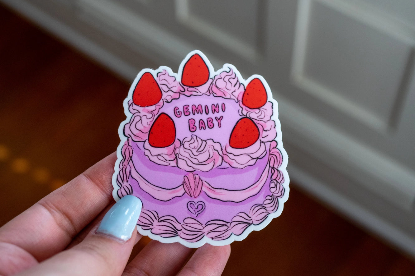 Sticker of a pink cake with red strawberries and 'Gemini Baby' text held by a hand.