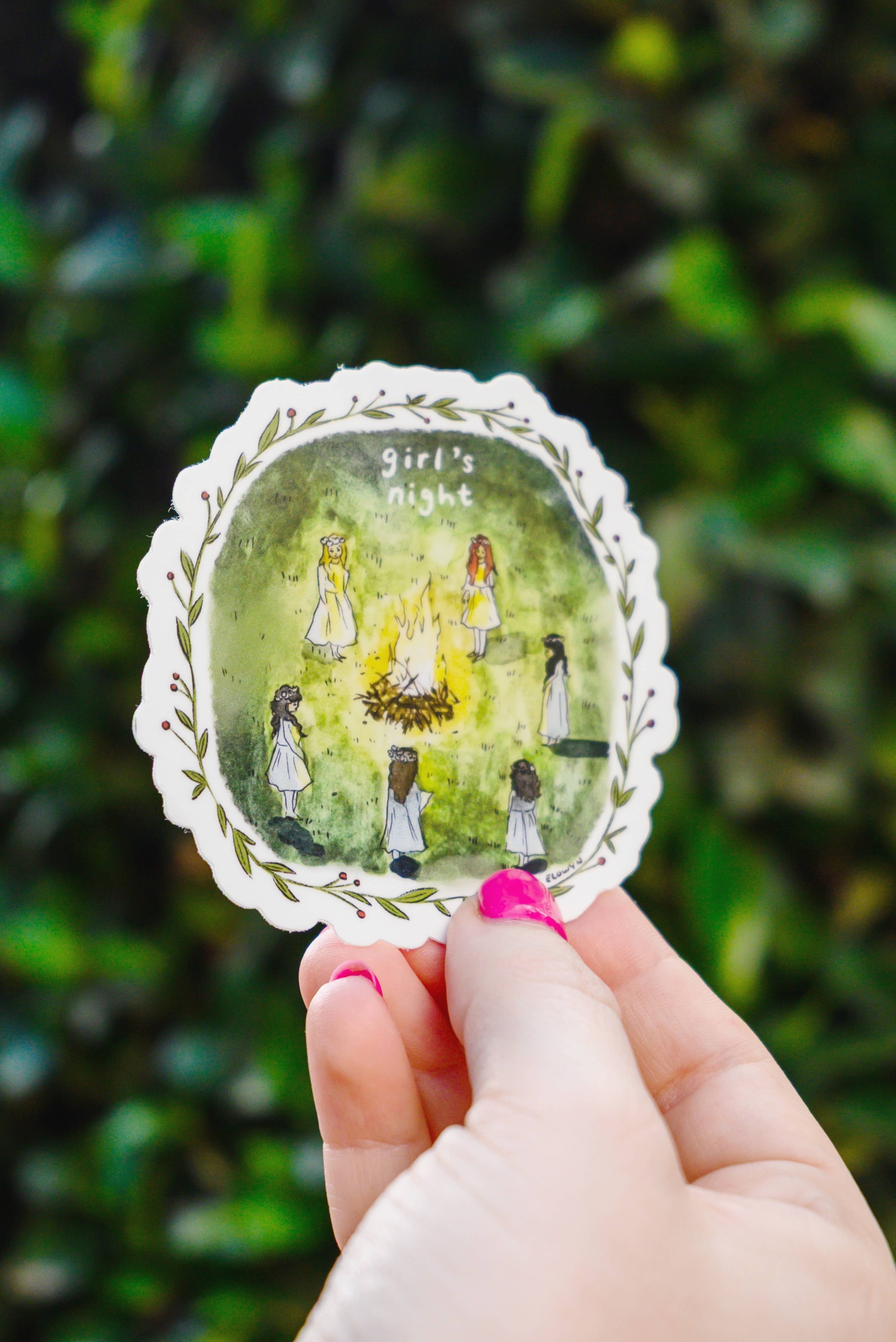 Hand holding a decorative sticker with illustrations of women and text 'girl's night' against a green leafy background.