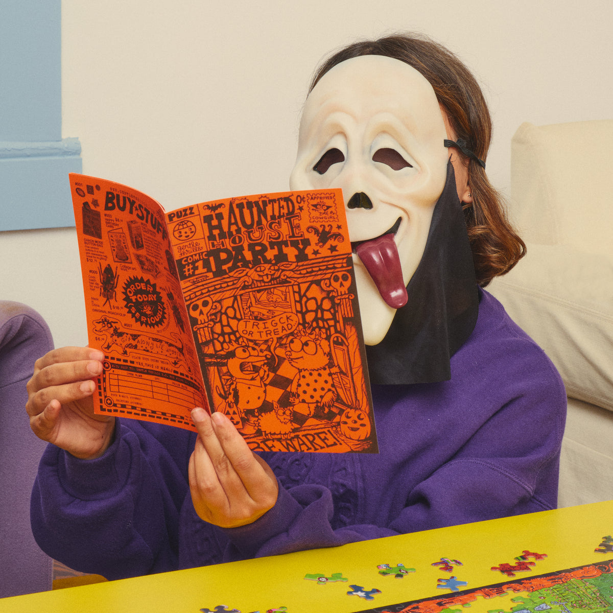 Person in a ghost mask and purple jumper reading the orange Haunted House Party comic booklet.