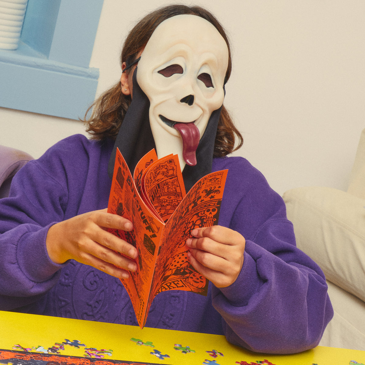 Person in a ghost mask unfolding the orange Haunted House Party booklet beside puzzle pieces.