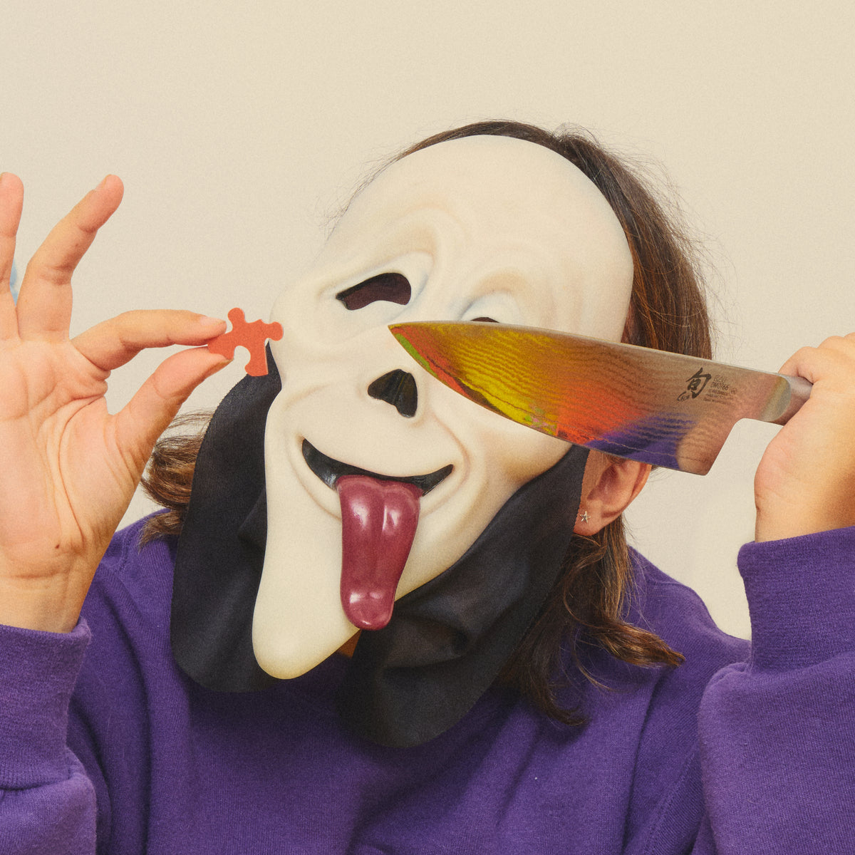 Person in a ghost mask holding an orange puzzle piece and a rainbow-coloured knife.