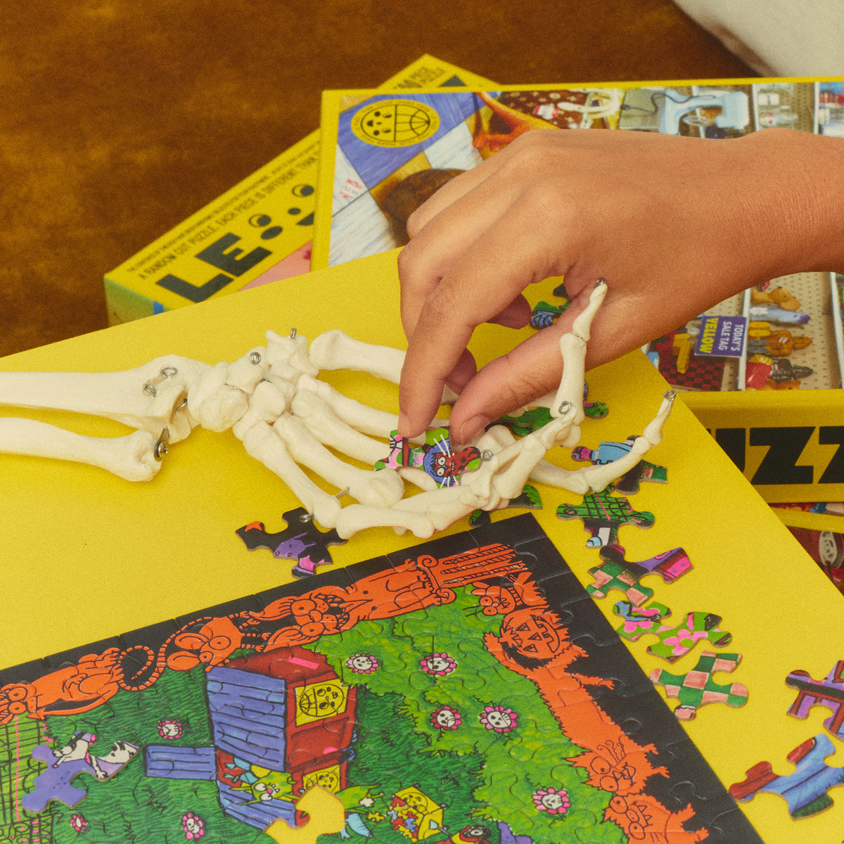 Hand picking up a Haunted House Party puzzle piece beside a skeleton hand prop on a yellow table.