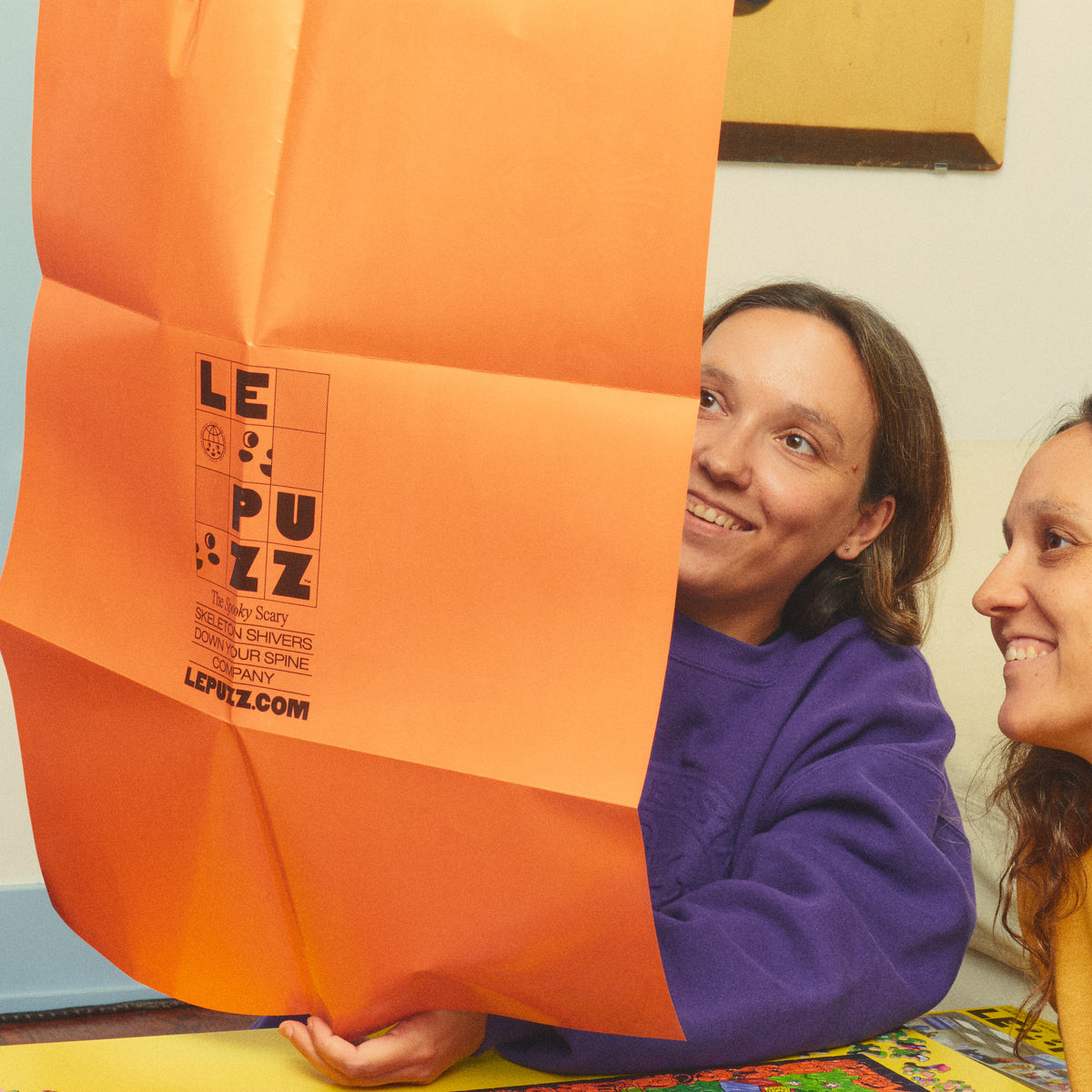 Two smiling people looking at the unfolded orange Le Puzz poster beside the Haunted House Party puzzle.