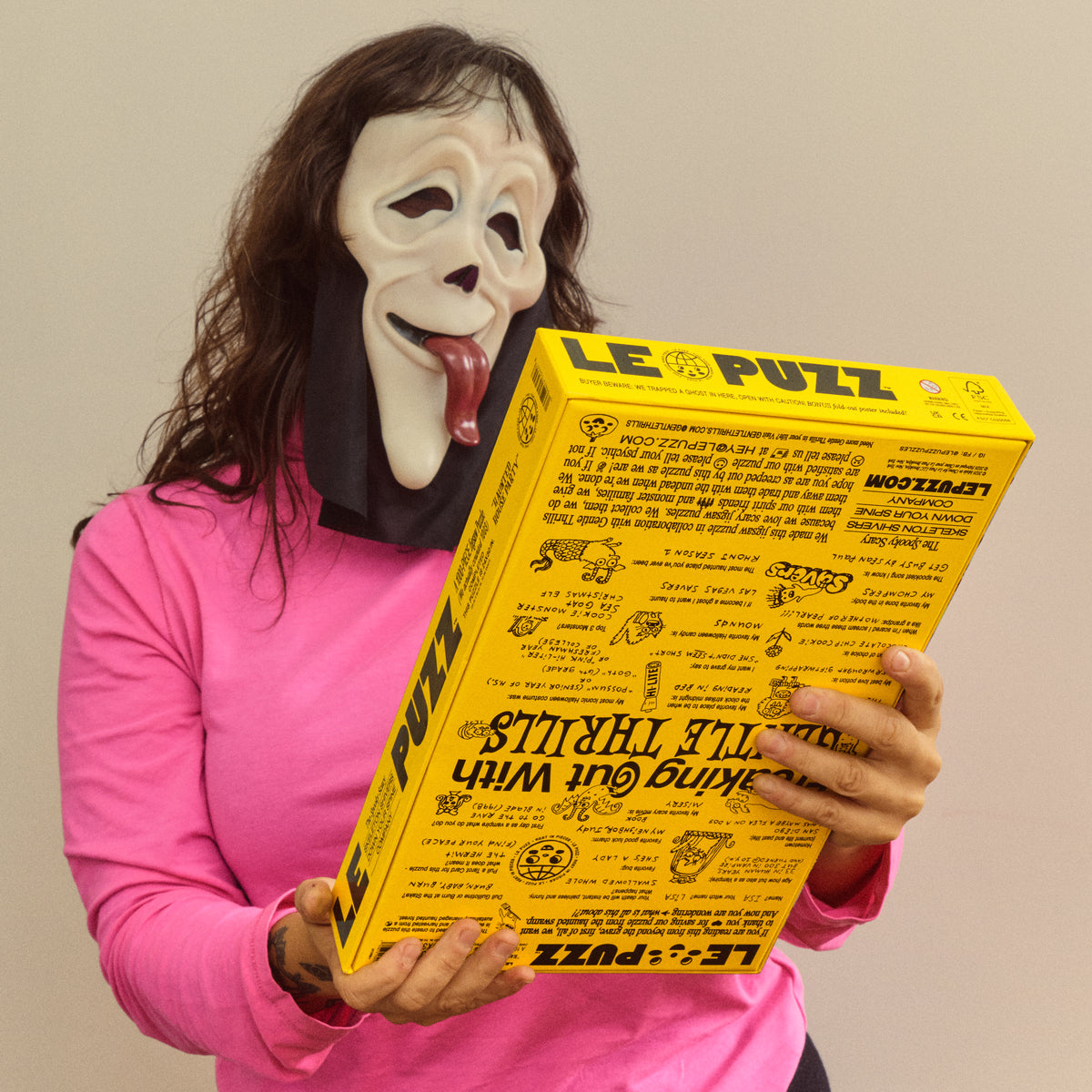 Person in a ghost mask and pink top holding the Haunted House Party puzzle box with its back facing forward.
