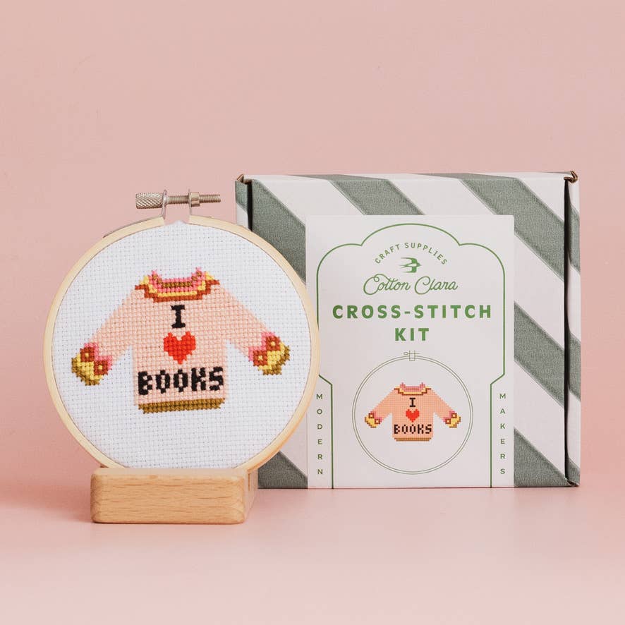 Cross-stitch kit with a completed design of a sweater saying 'I love books' next to its packaging on a pink background.
