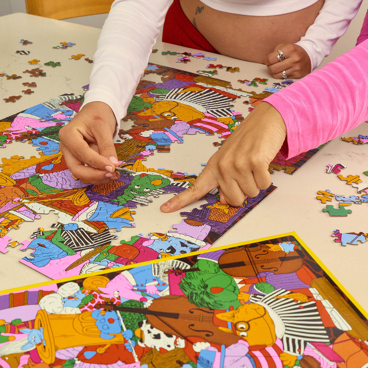 Two people assembling the colourful Le Puzz K9 Symphony jigsaw puzzle, with a matching reference image in the foreground