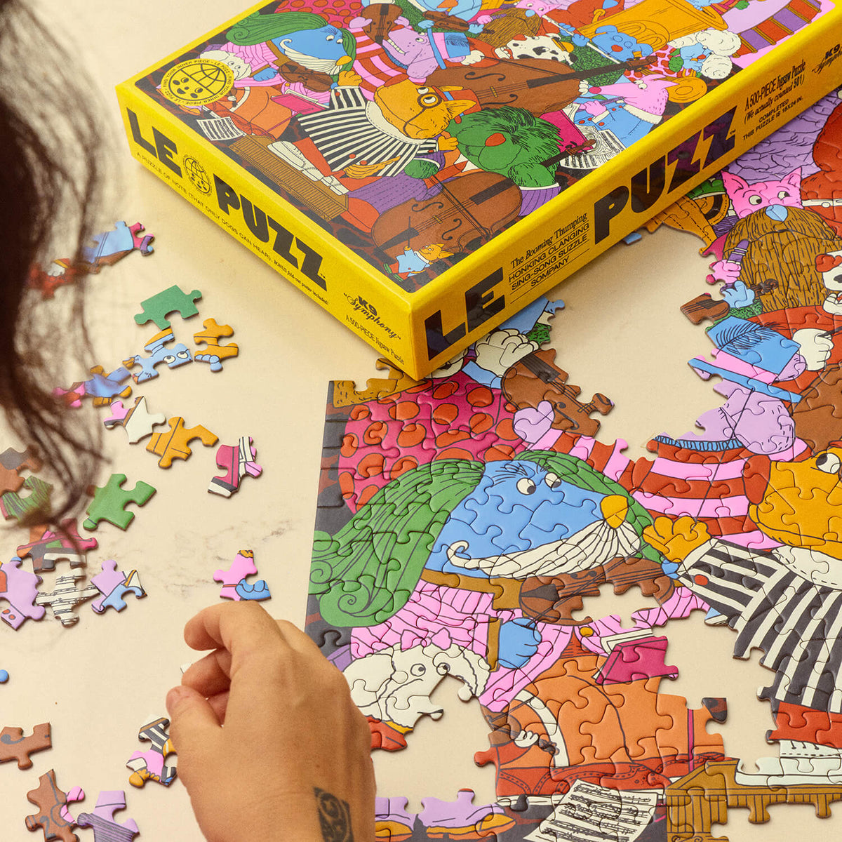 Le Puzz K9 Symphony yellow puzzle box beside a partially assembled jigsaw featuring colourful dogs playing instruments.