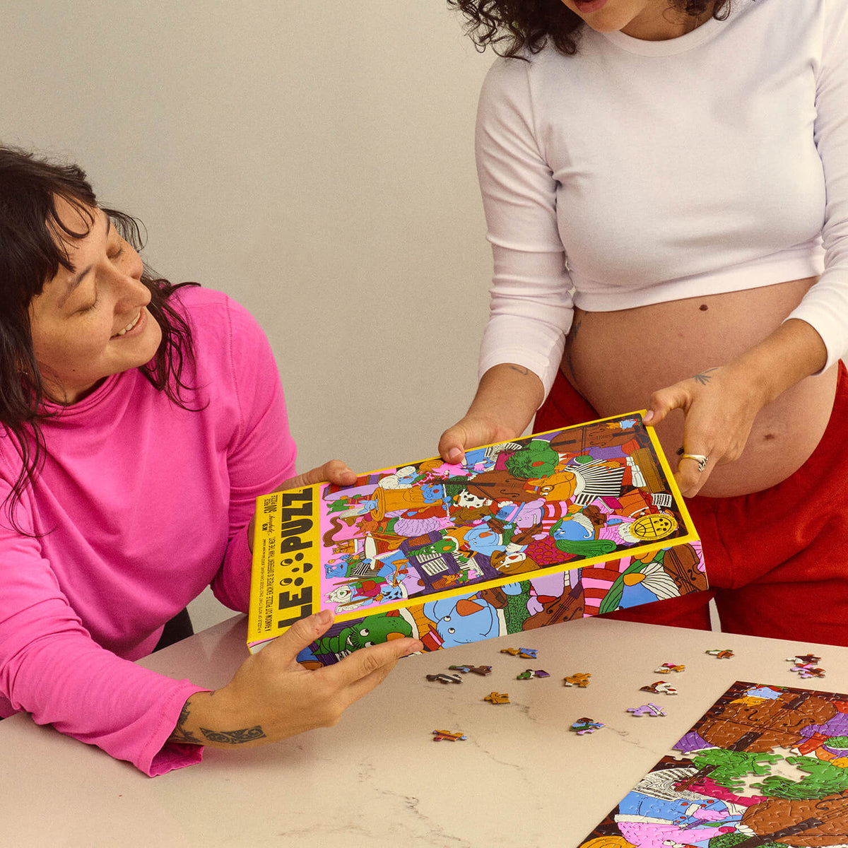 Two people holding the yellow Le Puzz K9 Symphony puzzle box above a table with loose pieces and a partially assembled puzzle.