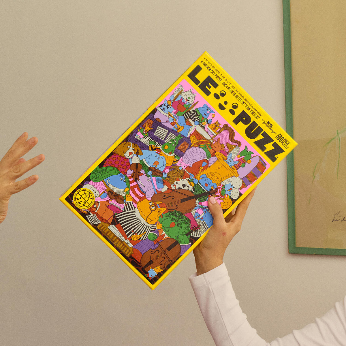 Hand holding the yellow Le Puzz K9 Symphony 500-piece puzzle box at an angle, showing colourful dogs playing instruments.