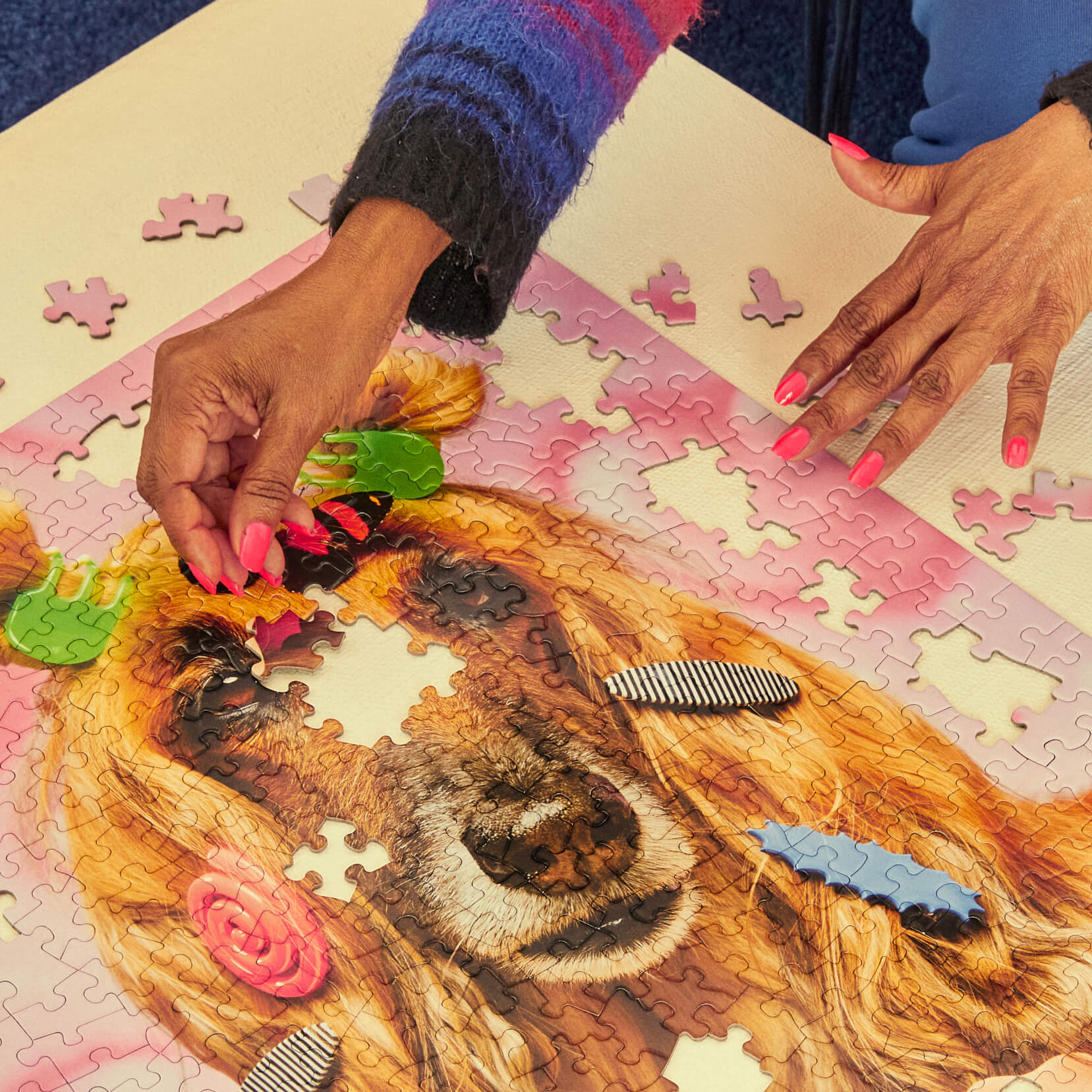 Person assembling a puzzle of a dog with flowers, wearing a blue sweater and pink nail polish.