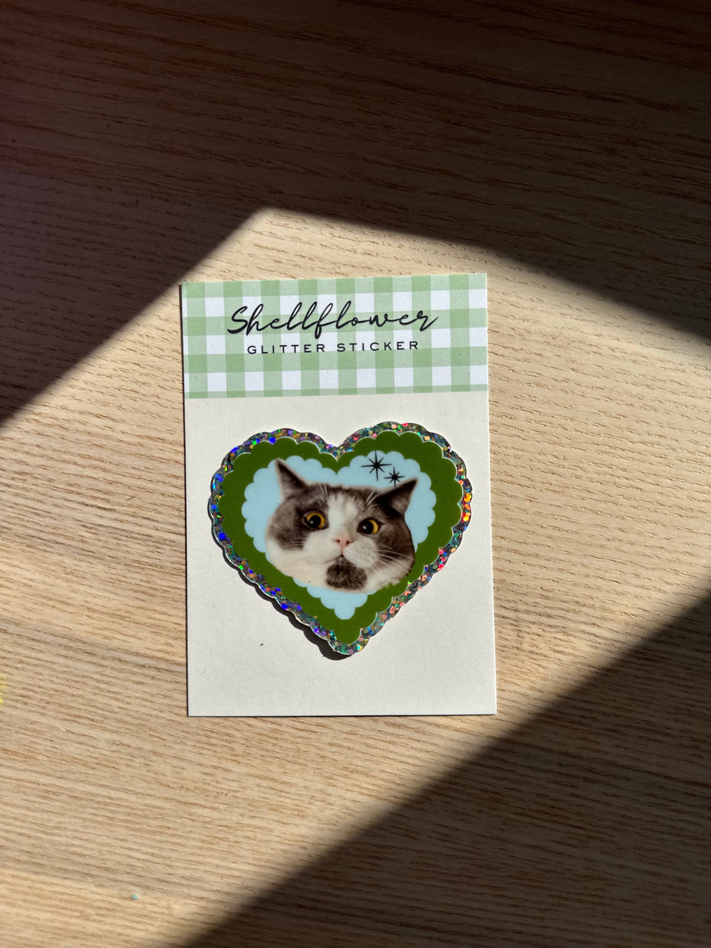 Heart-shaped sticker of a cat on a card with 'Shellflower' branding, placed on a wooden surface.
