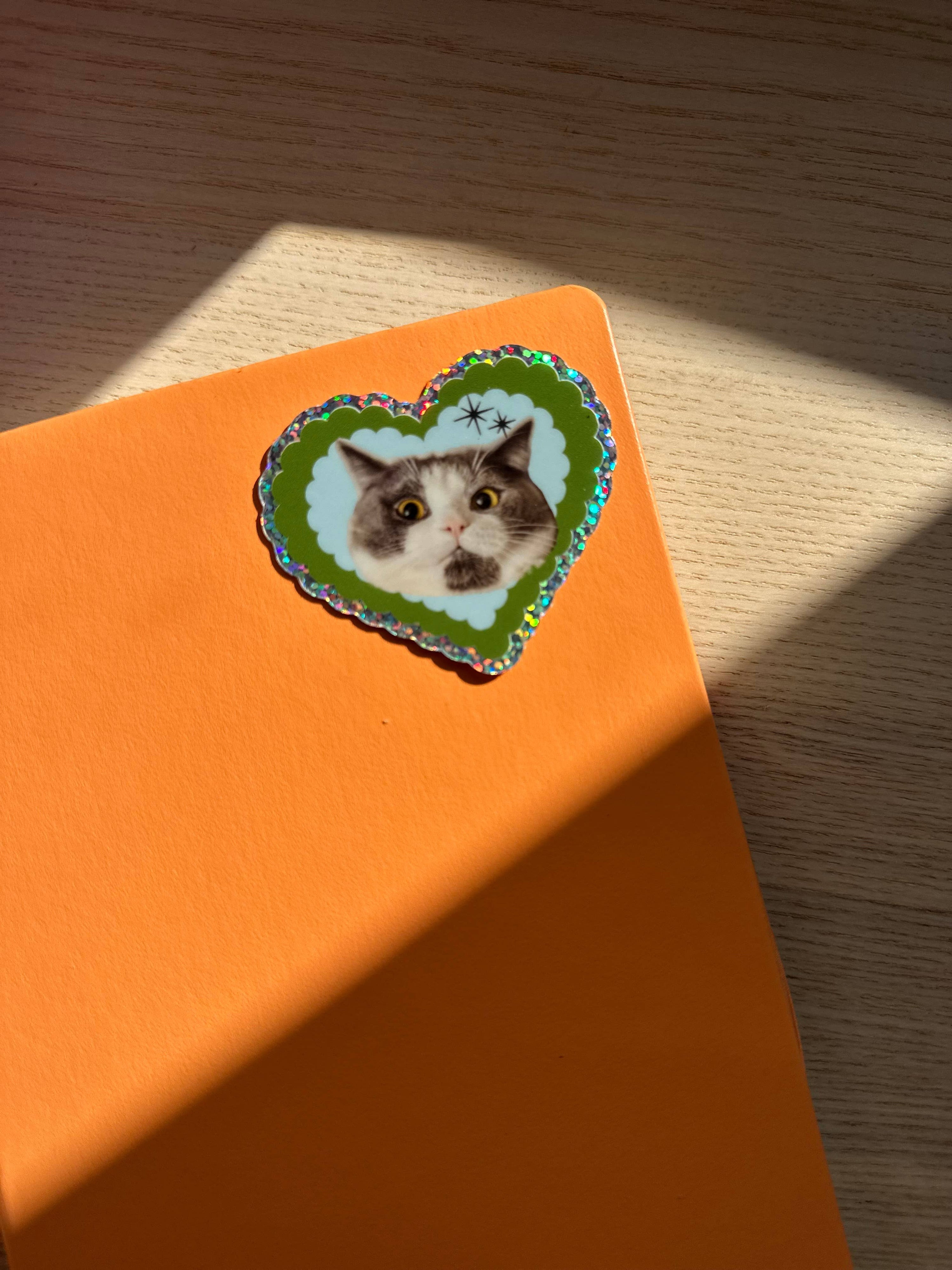 Heart-shaped sticker of a cat on an orange surface with sunlight casting shadows.