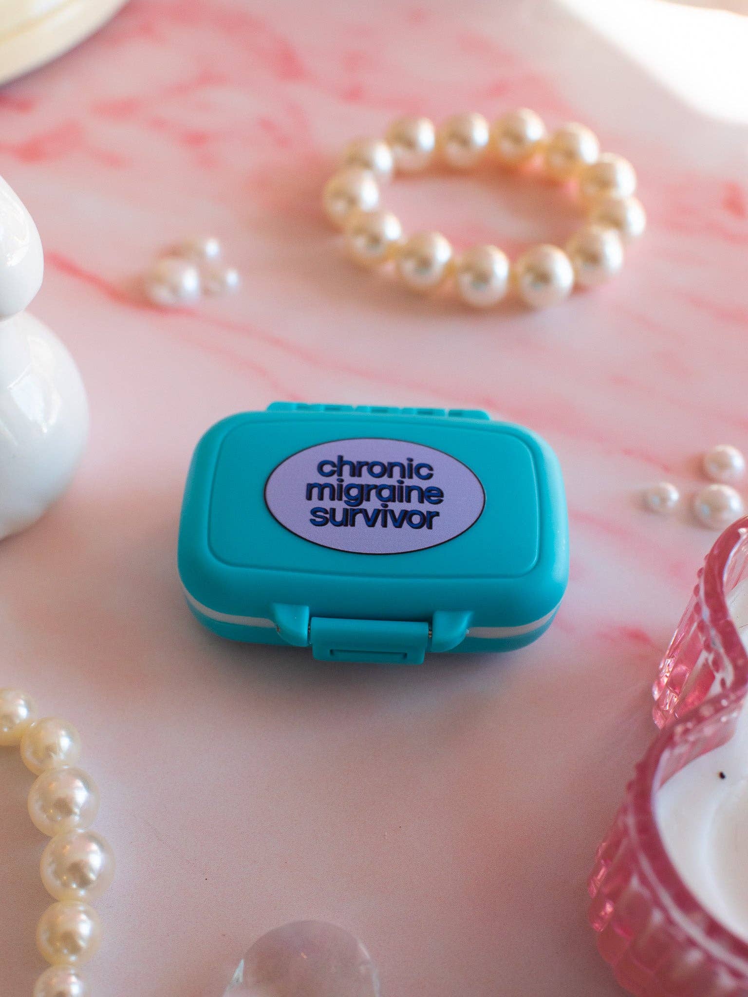 Small teal box with a purple label reading 'chronic migraine survivor' on a pink surface with pearls.
