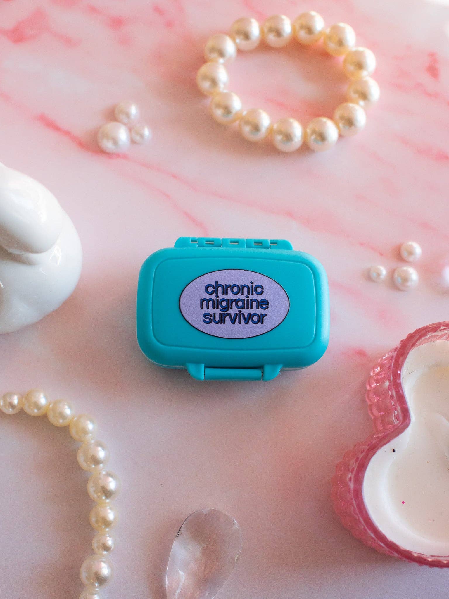 Teal pill box with 'chronic migraine survivor' text on a pink marble surface with pearls.
