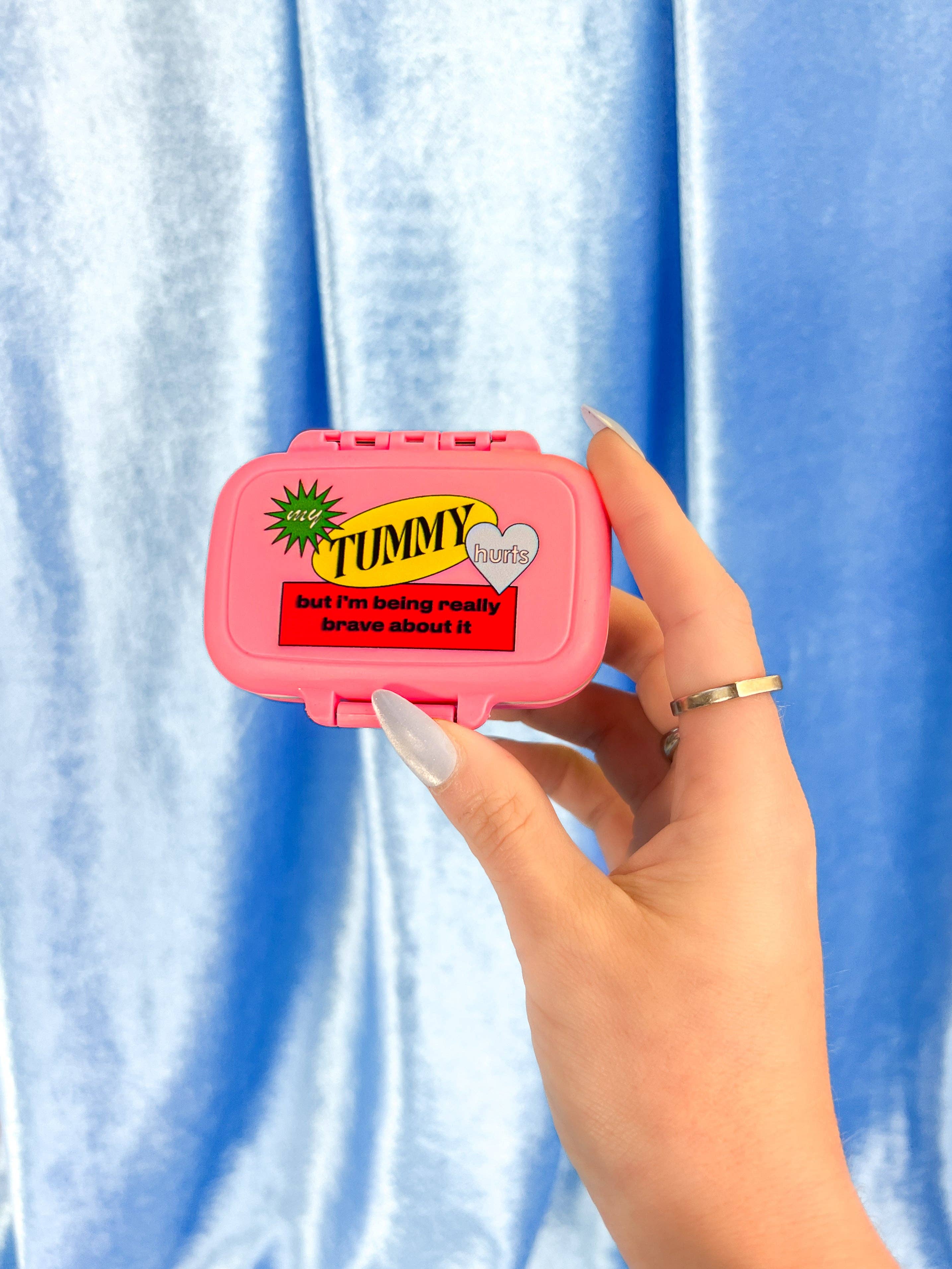 Pink pill box with a humorous label held by a hand against a blue curtain background