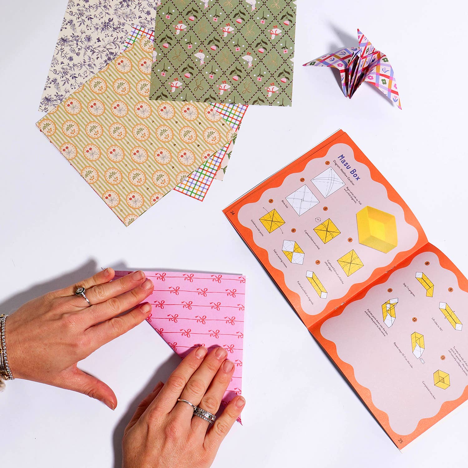 Hands holding a pink patterned paper with an open box of stationery items on a white background