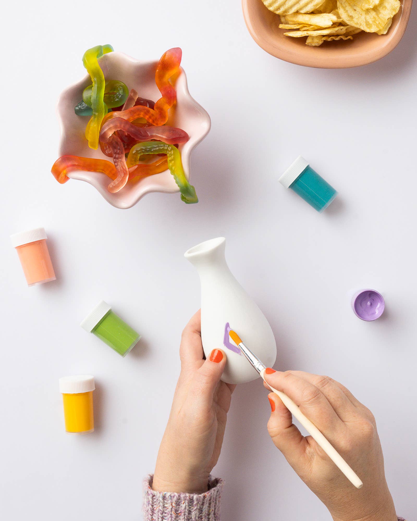 Person painting a ceramic vase with colorful glazes on a white surface, surrounded by gummy bears and chips.